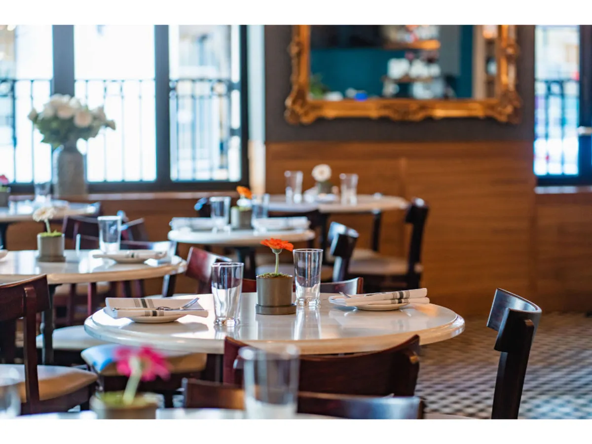 A cozy, elegant restaurant setting with marble tables, vibrant flower centerpieces, and wooden chairs. The decorative mirror adds depth, and the overall ambiance feels warm, social, and welcoming—ideal for families or groups sharing a meal.