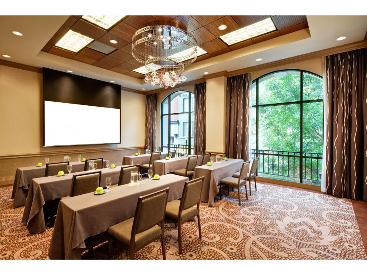 A spacious, well-lit room featuring long tables covered with crisp white tablecloths, a sparkling chandelier overhead, and a large window revealing lush greenery outside. A presentation screen is set up, suggesting active use for business meetings or workshops, while the natural light and outside view add a sense of openness and connection to the city.