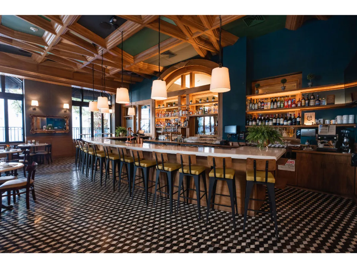 A warm and welcoming view of the bar area, featuring a prominent counter framed by hanging light fixtures, shelves filled with bottles, and a distinctive checkerboard floor. The ambiance suggests lively conversations over drinks and celebratory moments.