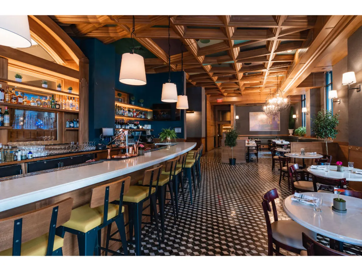 This image features a sophisticated yet relaxed bar area adorned with chandeliers, a decorative ceiling, and inviting dining tables. The scene suggests a lively social environment perfect for enjoying craft cocktails and seasonal small plates, with a vibrant ambiance that hints at urban nightlife and gathering.