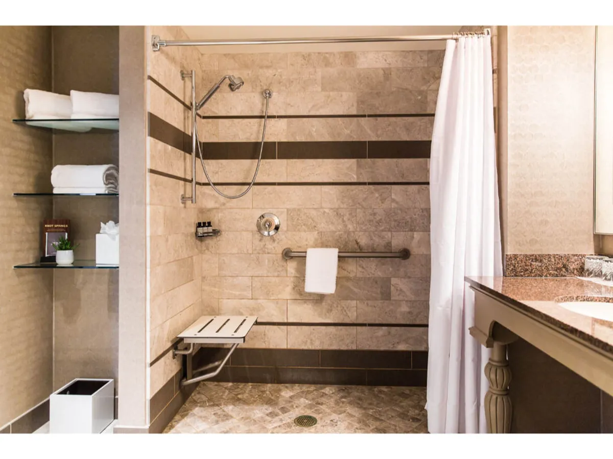 A serene bathroom scene featuring a spacious, modern shower area designed with accessibility in mind. The shower has a low threshold, making it easy to roll in, and there are supportive grab bars visible. Stylish shelves are neatly stacked with plush towels, and the granite countertop suggests upscale finishes. The overall ambiance is calm and spa-inspired, inviting relaxation and self-care.