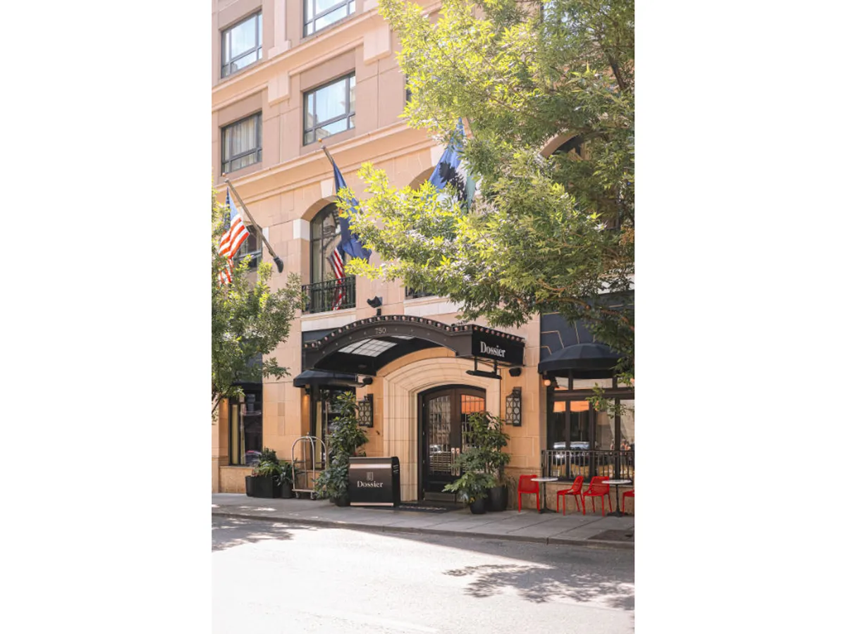 A street-level view of the hotel's exterior, featuring an inviting archway, outdoor seating area, flags, and lush greenery. The lively patio scene hints at barrier-free outdoor enjoyment and social gatherings.