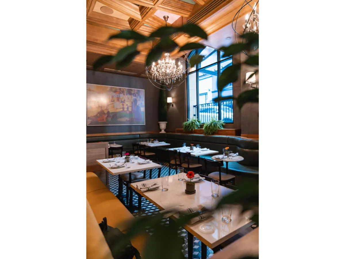 A sophisticated dining space with a chandelier, contemporary artwork, and plants in the background. The setting offers cozy tables that invite guests to relax, savor a meal, and enjoy a sense of comfort.