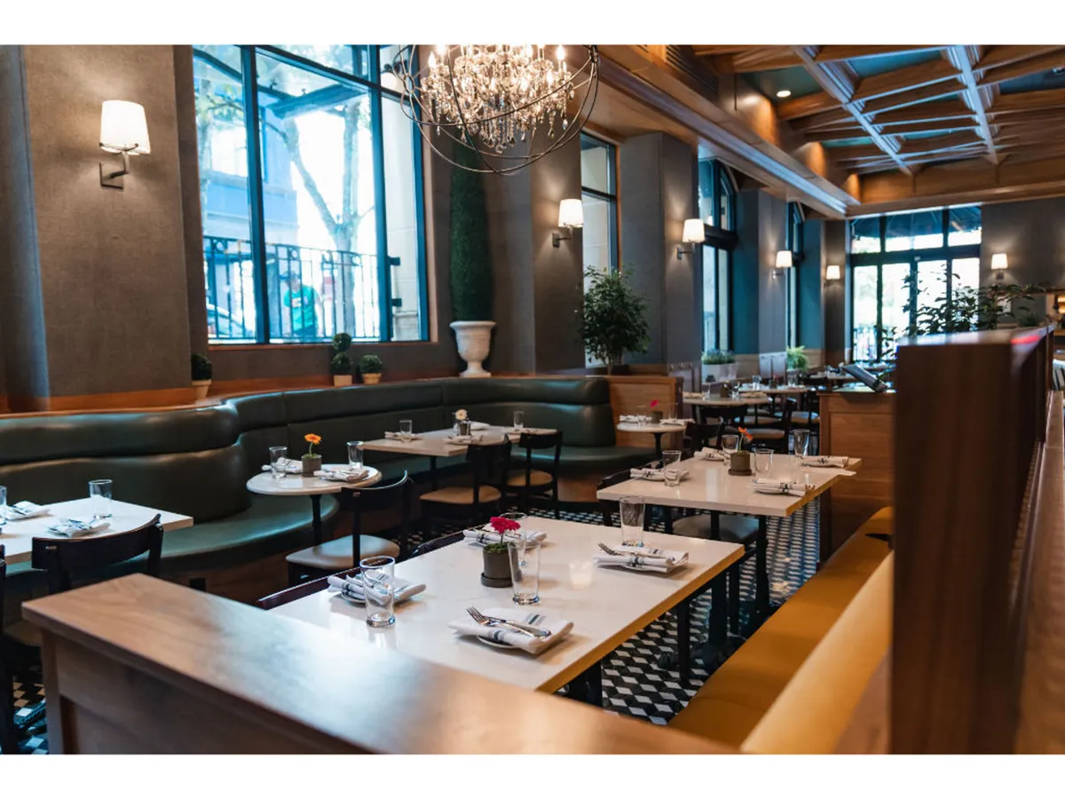 This interior scene features a bright, elegant dining area with a striking chandelier, lush greenery, and tables set with cutlery. The room emanates a welcoming and social atmosphere, with subtle nods to natural elements that suggest a connection to the outdoors.