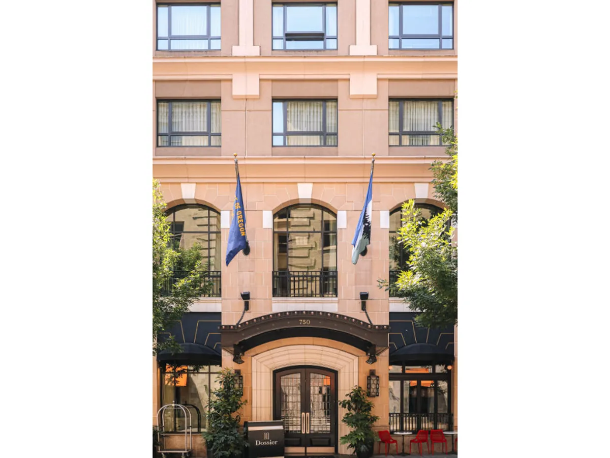 The hotel's main exterior with a broad entrance, prominent flags, large windows, and a decorative archway. The scene captures an inviting, open setting that sits comfortably within its urban context, ideal for guests coming and going on city explorations.