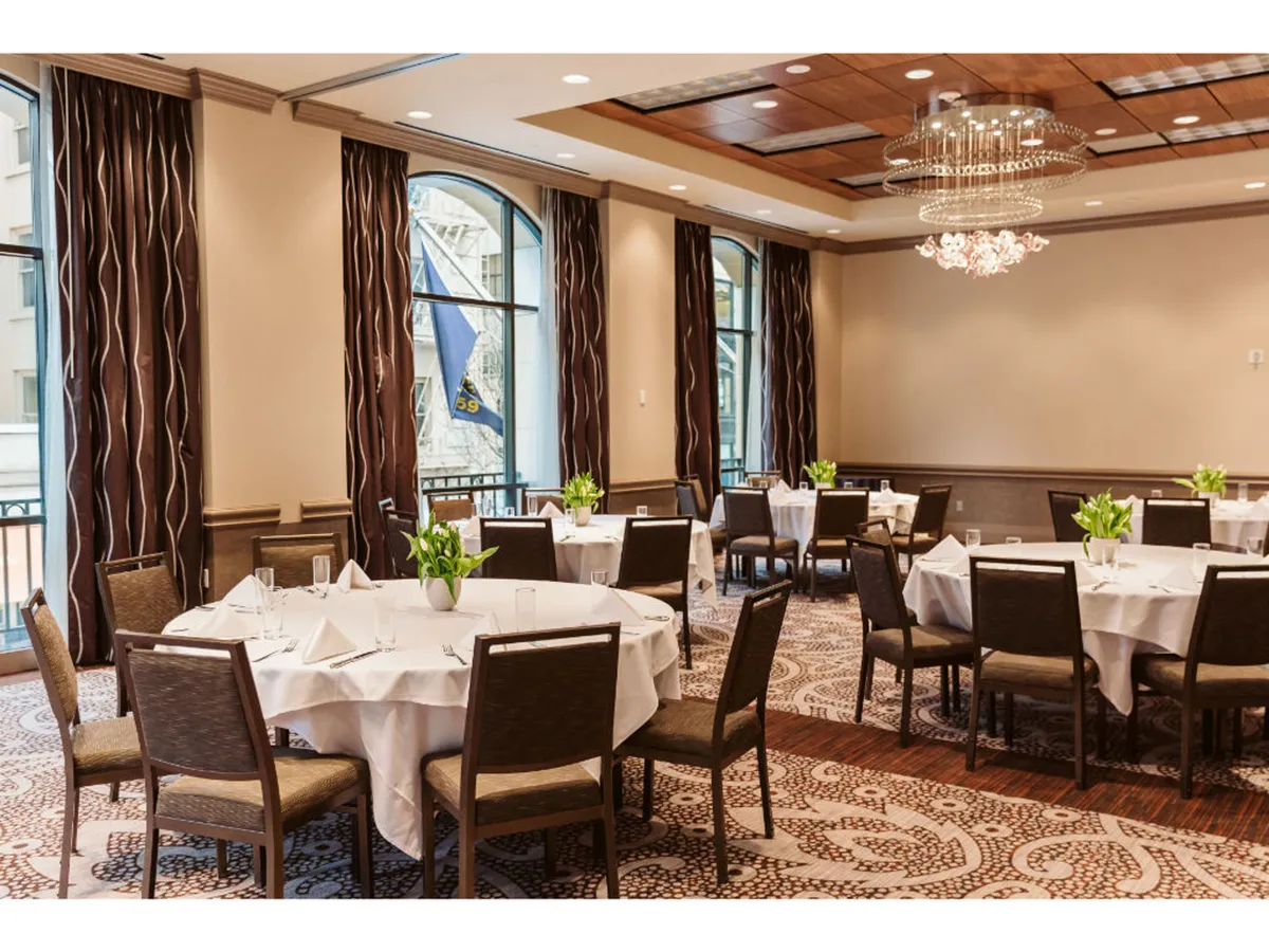 A refined dining area featuring round tables set for groups, a sparkling chandelier, and expansive windows draped with curtains. Daylight streams through, offering a glimpse of greenery outside and creating a welcoming space suitable for gatherings.