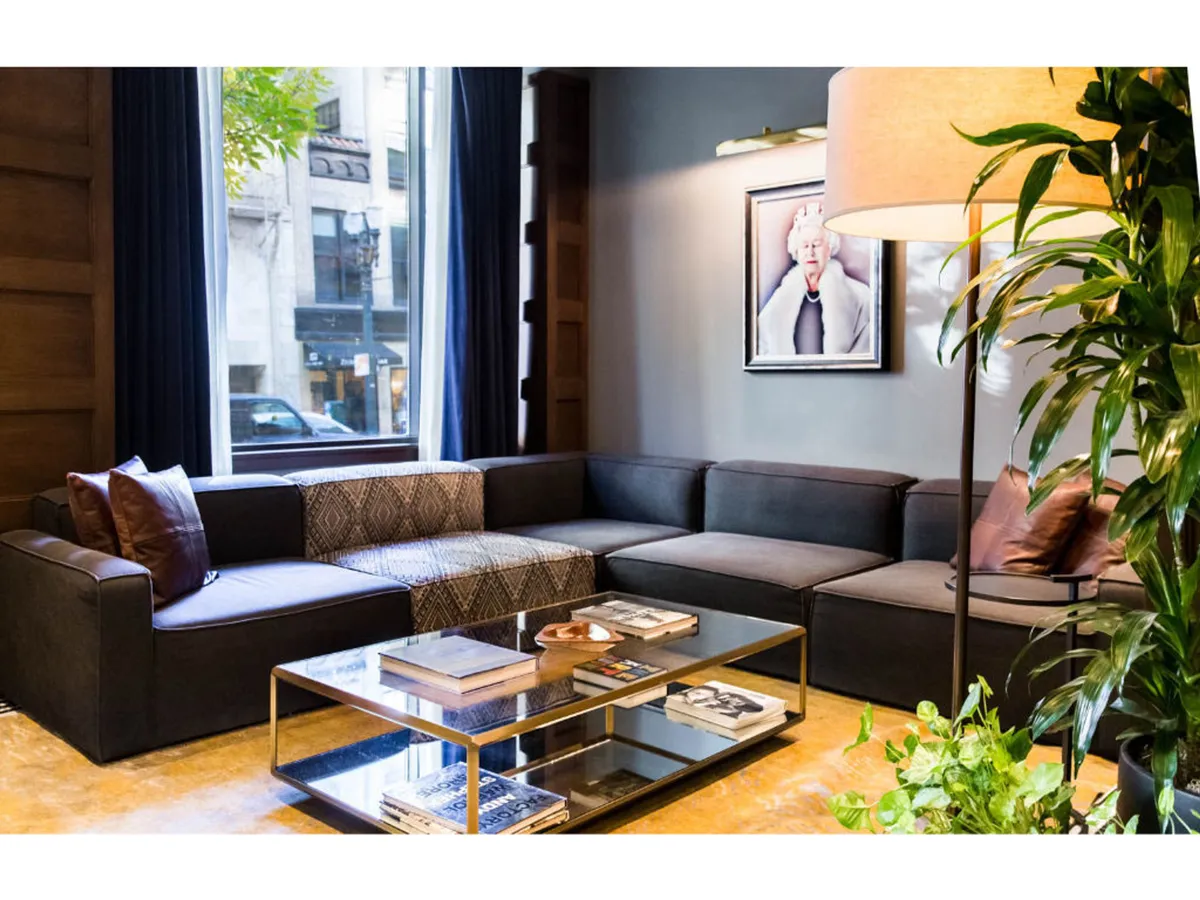 A stylish living area featuring a large sectional sofa, glass coffee table, contemporary artwork on the wall, a floor lamp, and lush indoor plants. The overall feel is cozy and sophisticated, ideal for both relaxation and light work.
