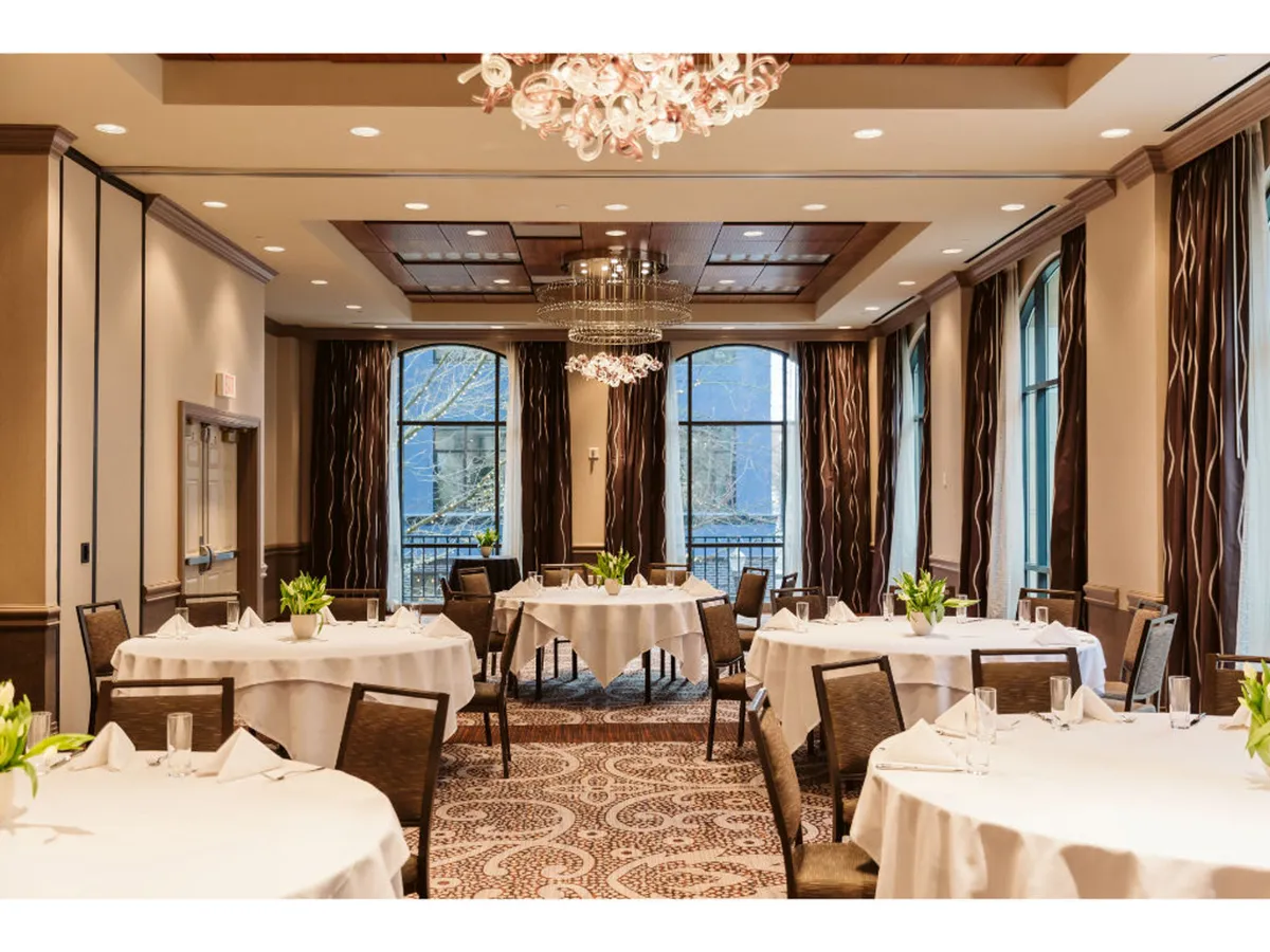 The image shows an elegant dining room with large windows draped in fabric, letting in natural light and offering a glimpse of the city. White tablecloths, a striking chandelier, and tasteful wall art create an atmosphere of refined urban dining.