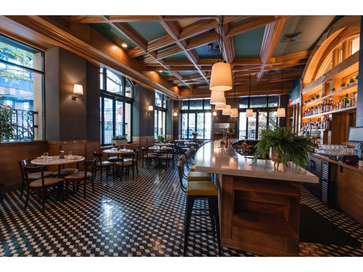 This image showcases Bar Alder, featuring a stylish bar counter, dining tables, and large windows that flood the space with natural light. The setting is lively, with guests enjoying signature cocktails at the bar and sharing meals while observing the vibrant street scene outside.
